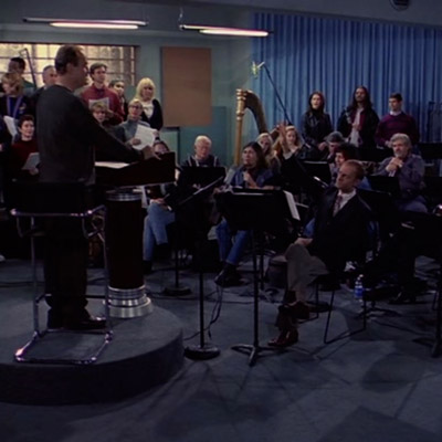 Fraiser in the position as the conductor. He is standing in front of the entire orchestra answering their questions.